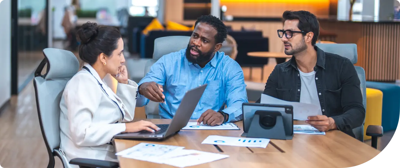 Three coworkers engage in a conversation at a conference table, surrounded by laptops, papers, and charts in a modern office.