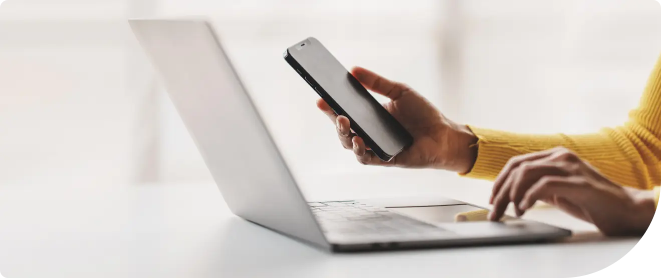 Person in a yellow sweater using a laptop while holding a smartphone, seated at a white desk in a bright room