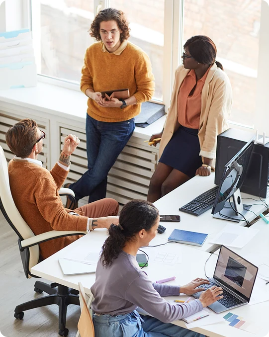 Four colleagues discuss a project around a desk with laptops and documents in a modern office