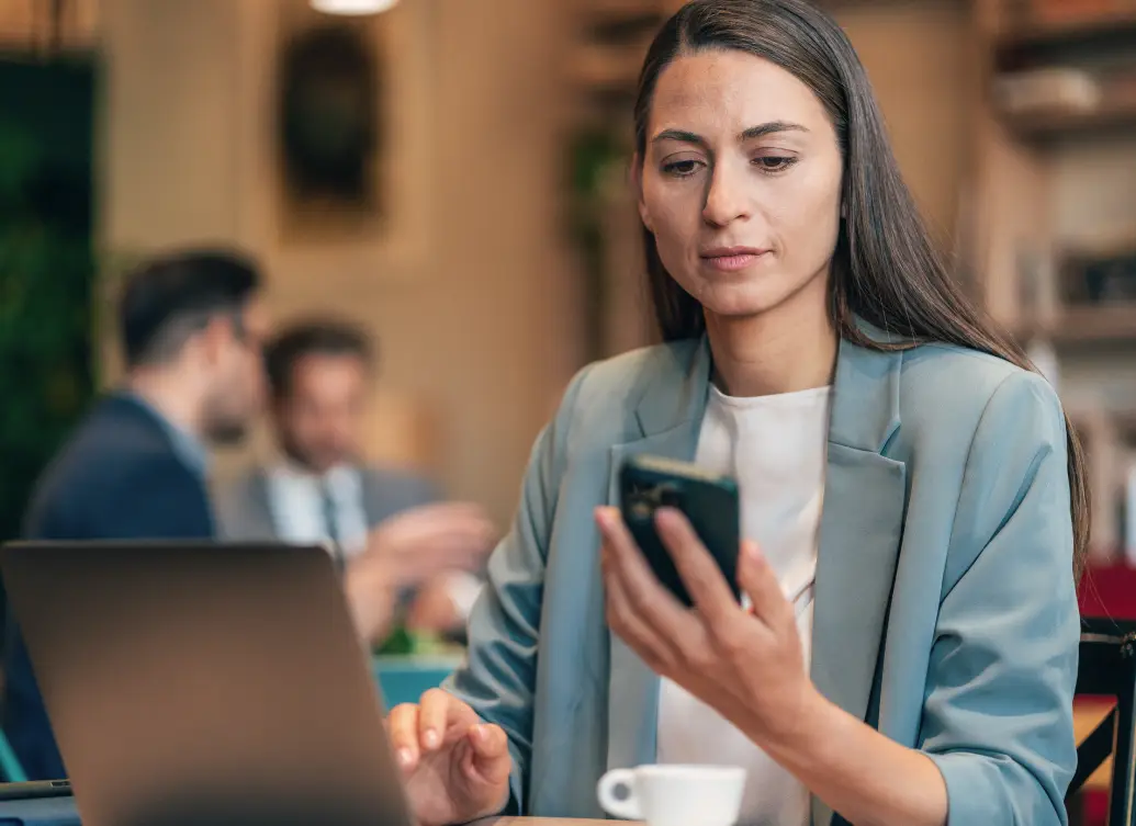 Woman in blazer sits at a desk with laptop, looking at her phone while two people talk in the background.