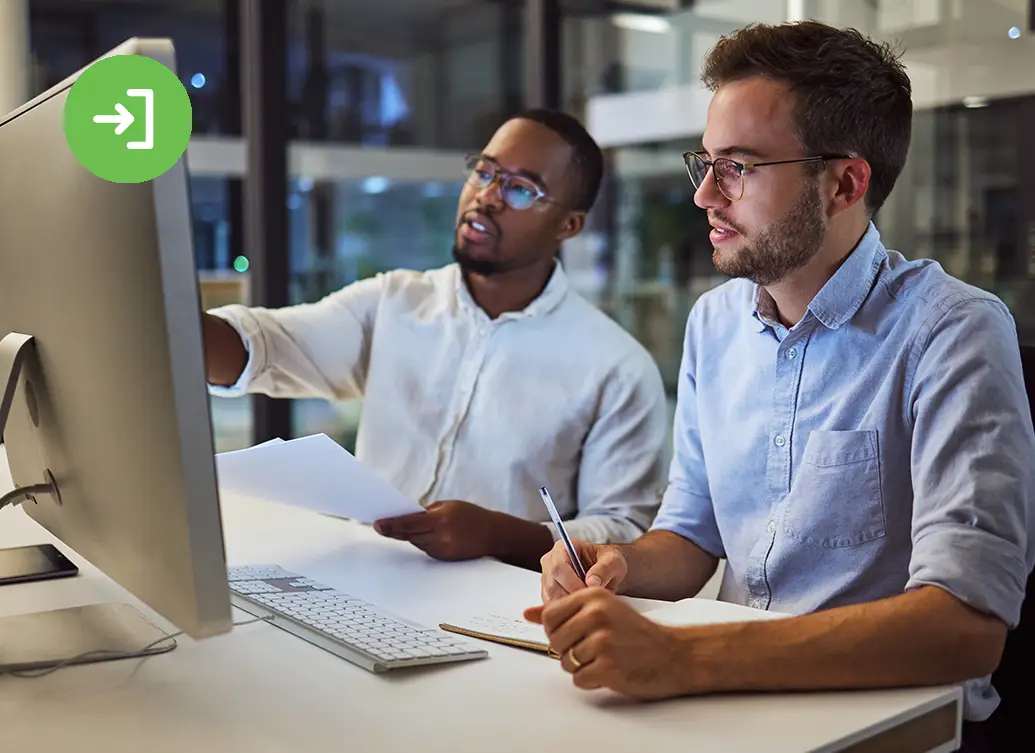 Two colleagues in an office work at a computer, one pointing at the monitor while the other takes notes with a pen and notebook.