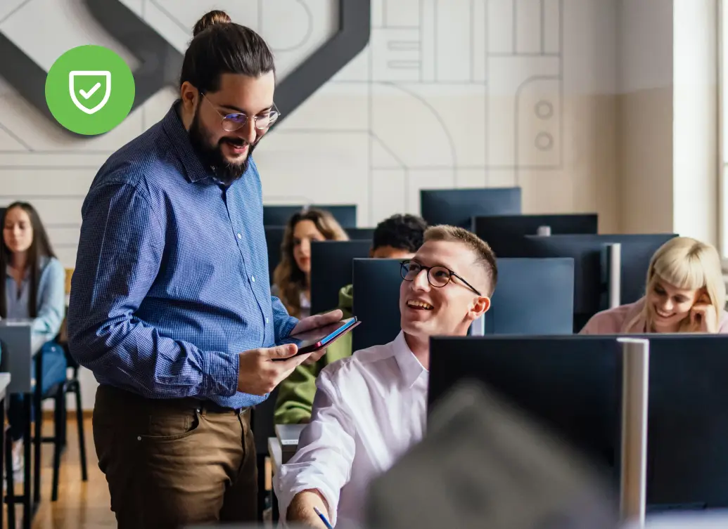 In an office, a man standing with a tablet speaks to a seated colleague who is smiling, while other people work at computers in the background.