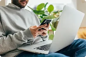 A person sitting indoors, looking at their smartphone with a laptop open on their lap.